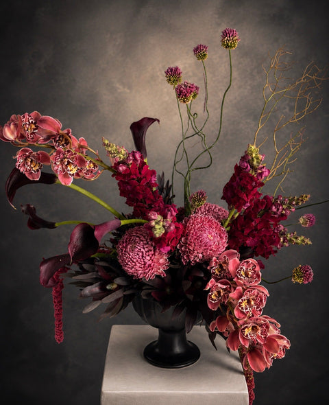 Floral arrangement with red and burgundy flowers in a black vase on a gray background, which include premium orchids, snapdragons and Calla Lilies