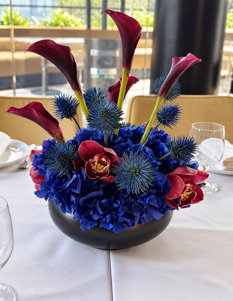 A vibrant floral centerpiece in a round black bowl, featuring royal blue hydrangeas, blue sea holly, burgundy calla lilies, and red orchids on a white tablecloth in a restaurant setting.