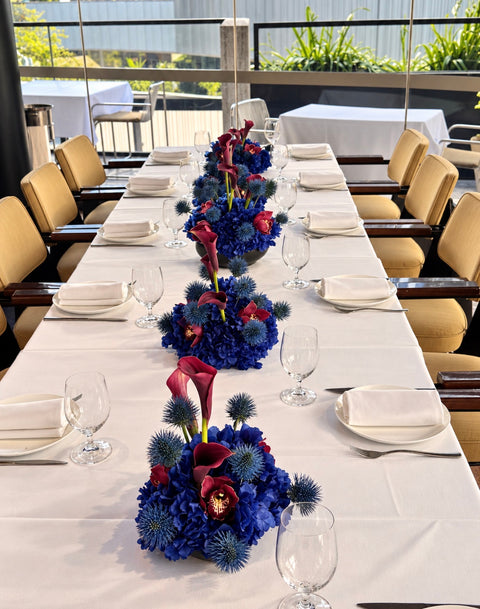 A long dining table set with white tablecloths, white plates, folded napkins, and clear wine glasses, featuring three blue floral centerpieces with blue hydrangeas, blue globe flowers, burgundy calla lilies, and red orchids in an indoor event space.
