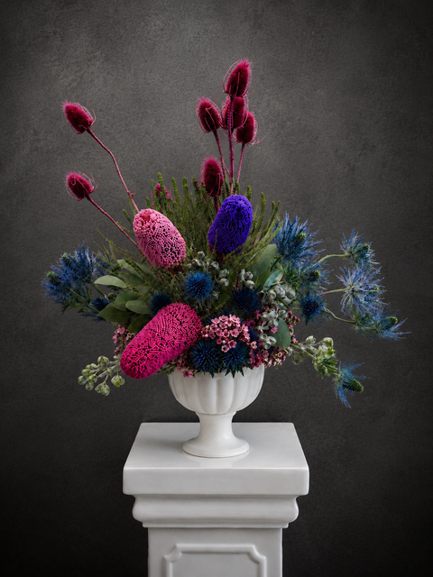 A vibrant native floral arrangement featuring magenta, blue, and purple wildflowers with green foliage, presented in a white classical urn vase atop a white pedestal against a dark textured background.
