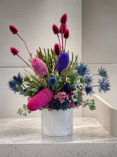 A native floral arrangement in a white marble vase, featuring vibrant magenta, blue, and purple flowers with green foliage, displayed on a marble surface against a textured light grey wall in an office setting.