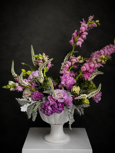 Luxury purple floral arrangement with snapdragons and lisianthus in a white ceramic pedestal vase against a dark background