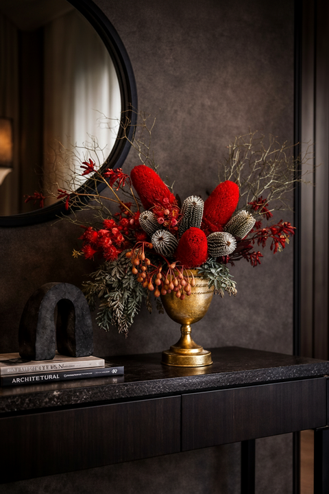 Australian native decorative floral arrangement with red flowers and greenery in a gold vase on a dark surface.