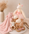 A soft plush unicorn toy in a pink tutu sits on a beige gift box, styled with a pink plush blanket, a small stuffed animal, wooden teething rings, and a neutral fabric backdrop.