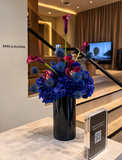 A floral arrangement in a tall black vase on a marble counter, featuring blue hydrangeas, burgundy calla lilies, and blue sea holly, with a Bang & Olufsen sign and brand plaque nearby.