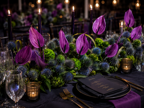 Decorative table setting with purple flowers, candles, and black plates.
