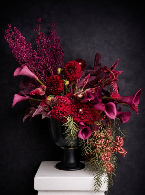 Moody crimson floral arrangement in a black vase on a white pedestal against a dark background
