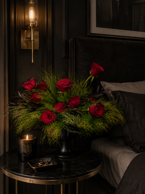 Decorative arrangement of red roses on a table next to a bed in a dimly lit room.