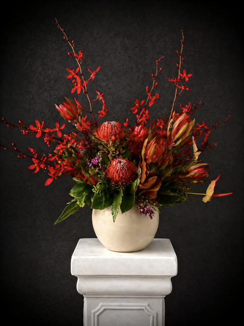 Decorative floral arrangement with red orchids and Natives in a beige vase on a white pedestal against a dark background