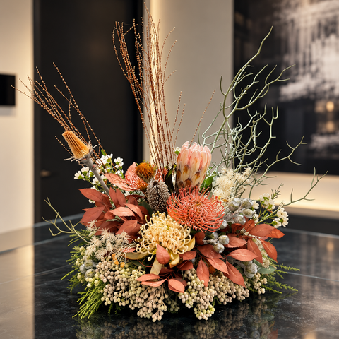 Luxury Australian native floral arrangement featuring blush protea, coral pincushion flowers, cream Grevillea, rust-toned foliage and sculptural branches in a stone vessel on a black marble surface, by Elysian & Bloom in Sydne
