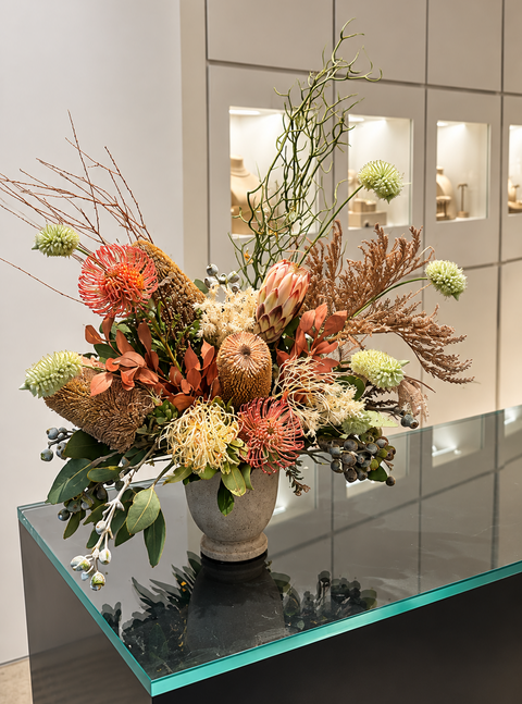 Luxury Australian native floral arrangement featuring blush protea, coral pincushion flowers, cream Grevillea, rust-toned foliage and sculptural branches in a stone vessel on a black marble surface, by Elysian & Bloom in Sydney