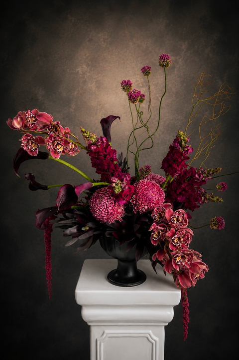 Moody Burgunduy Floral arrangement in a black vase on a white pedestal against a dark background with Cybidium orchids, snapdragons, amaranthys and Allums