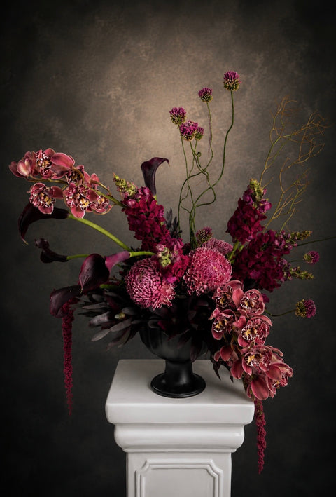 Floral arrangement in a black vase on a white pedestal against a dark background featuring a burgundy colour palette including orchids and snap dragons