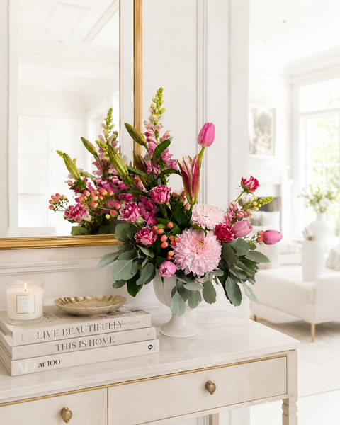 Pink Floral arrangement on a table with books and a candle in a bright room featuring disbuds, tulips, roses and snapdragons