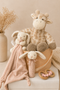A styled nursery gift set featuring a plush giraffe, a plush bunny, a soft pink security blanket, a wooden teething ring, and a wooden teething toy with pink beads, arranged in a basket on a white block with dried flowers in the background.