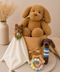 A styled baby gift set featuring a large caramel plush puppy toy sitting in a tan gift box, accompanied by a cream-colored plush blanket with a small brown teddy bear, a crocheted brown puppy toy with a blue and white striped scarf, and a wooden star bead teether.