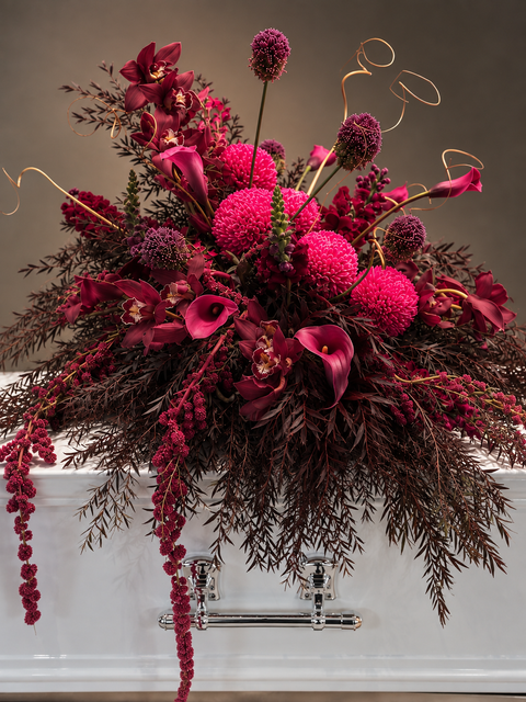 A dramatic Funeral Casket flower Cover in deep burgundy, magenta, and dark brown foliage, styled over a white bathtub with chrome faucets against a warm gray-brown background.