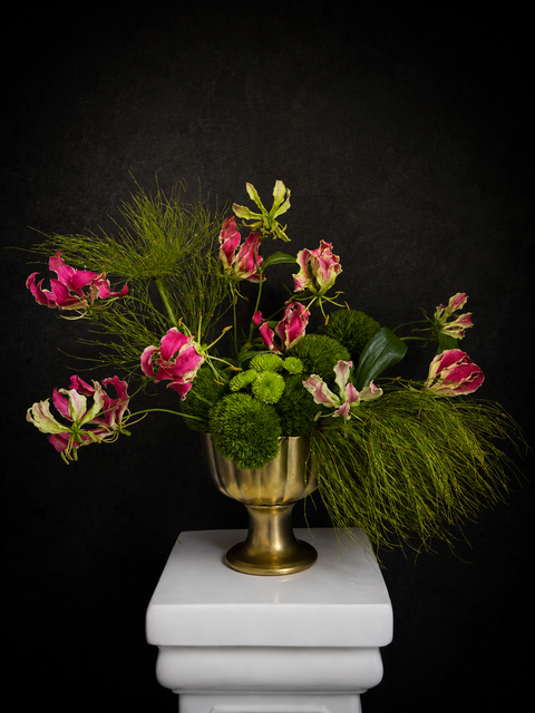 Luxury sculptural floral arrangement with pink gloriosa lilies, green chrysanthemums, and flowing foliage in a gold pedestal vase against a dark background