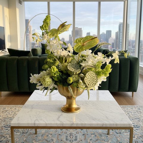 Green and white floral arrangement in a gold vase on a marble table with a cityscape view in the background