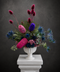 A vibrant native floral arrangement featuring magenta, blue, and purple wildflowers with green foliage, presented in a white classical urn vase atop a white pedestal against a dark textured background.